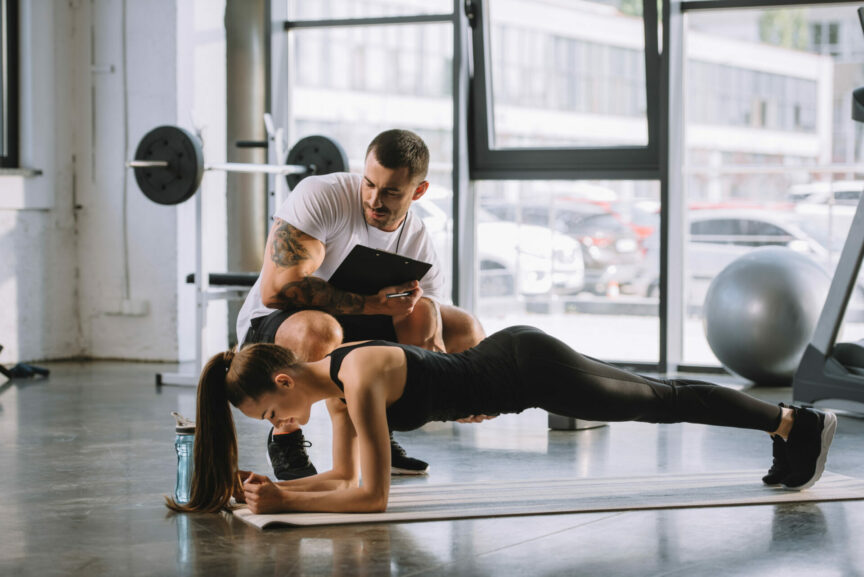 male personal trainer with clipboard and young athletic woman doing plank on fitness mat at gym
